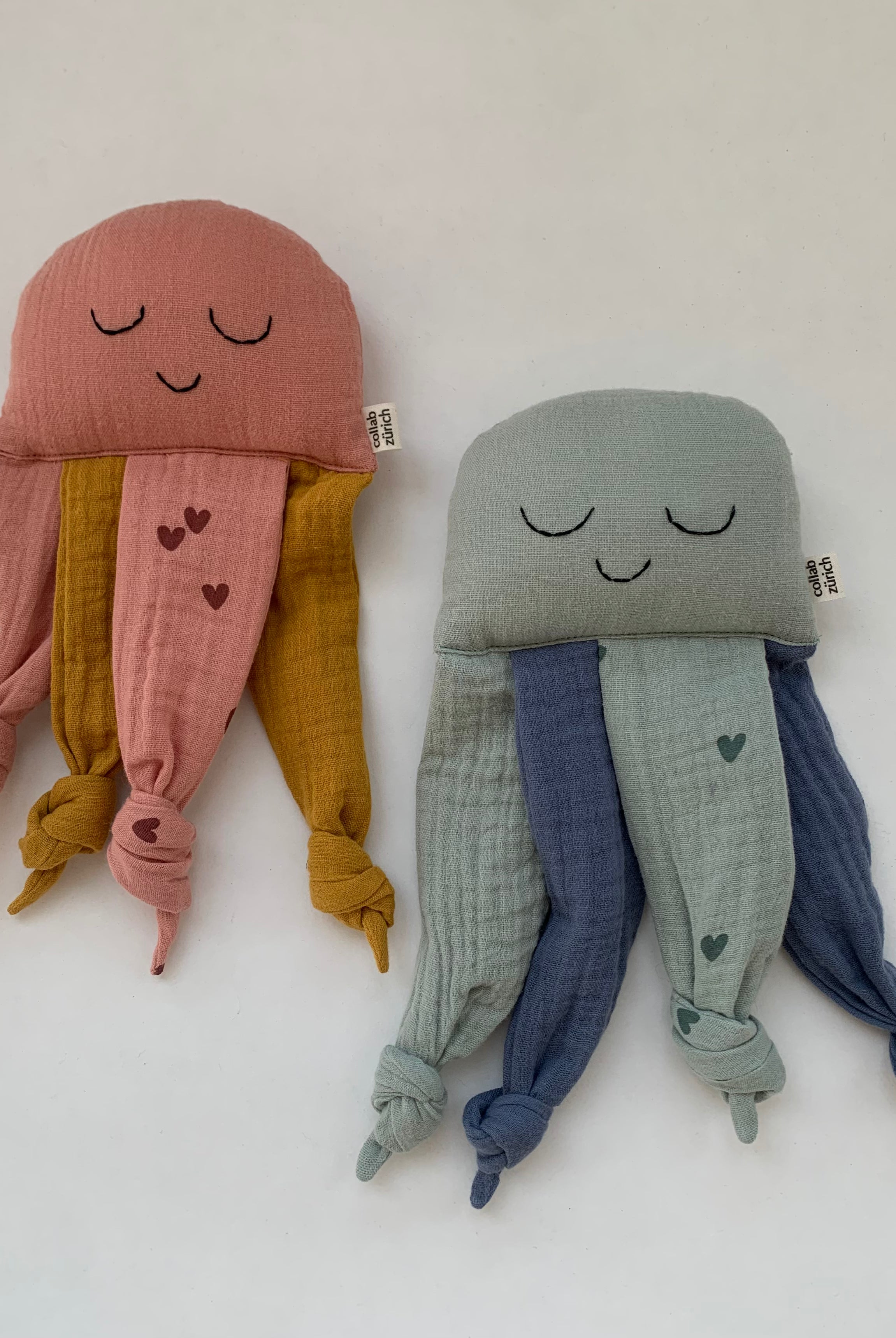 Two cute jellyfish-shaped muslin cloth toys on a plain white background. The jellyfish on the left is dusty pink with mustard yellow tentacles and small pink hearts.
