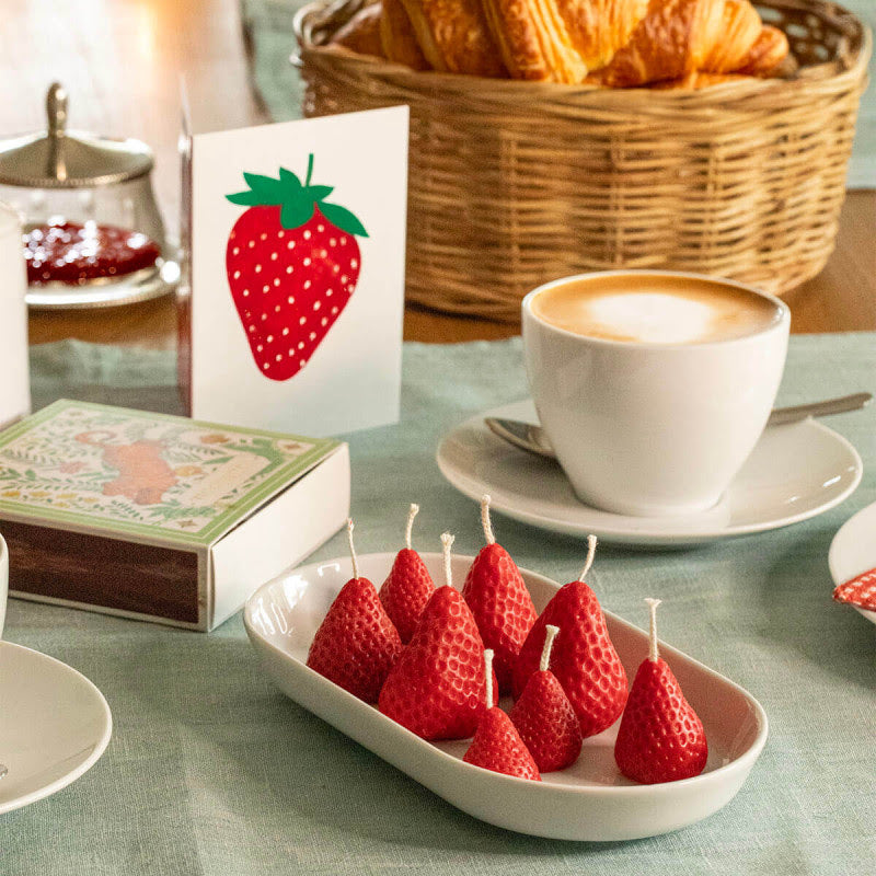 Table setting with strawberrie candles on a plate, a cup of coffee, and a basket of pastries.