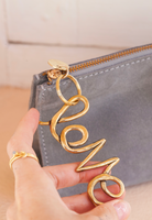 Gray clutch with gold 'Elo' zipper pull held by a hand.