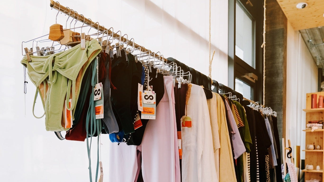 A clothing rack in a boutique displaying a variety of garments in different colors, including green, black, white, and pink, hanging from a rope-hung metal bar over a wooden bench.