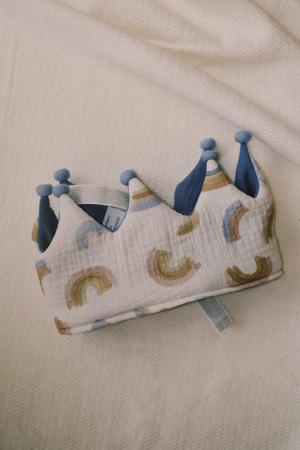 A handcrafted children's crown with a blue and multicolor pattern, featuring small pompons.