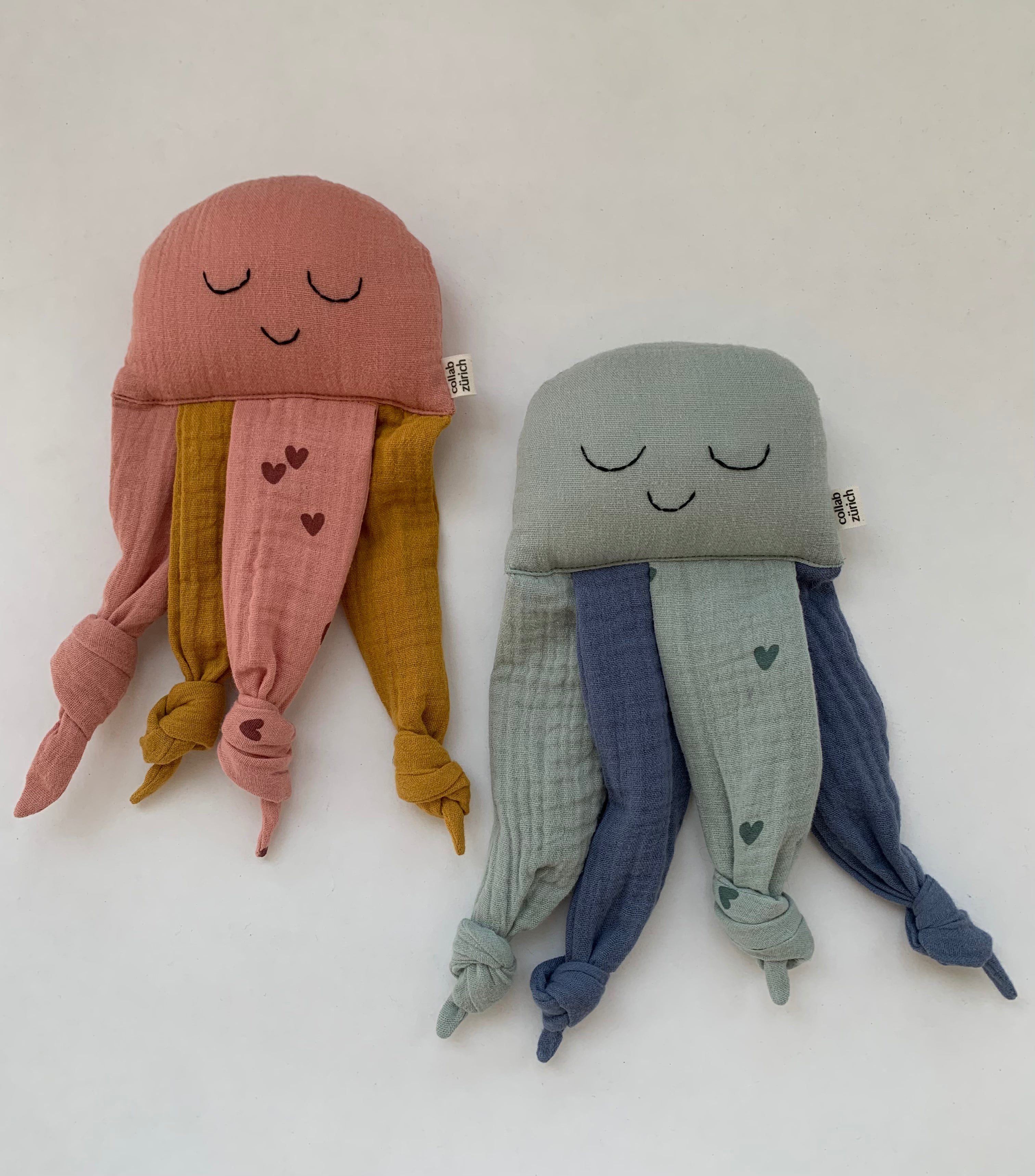 Two cute jellyfish-shaped muslin cloth toys on a plain white background. The jellyfish on the left is dusty pink with mustard yellow tentacles and small pink hearts.