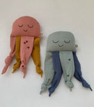Two cute jellyfish-shaped muslin cloth toys on a plain white background. The jellyfish on the left is dusty pink with mustard yellow tentacles and small pink hearts.